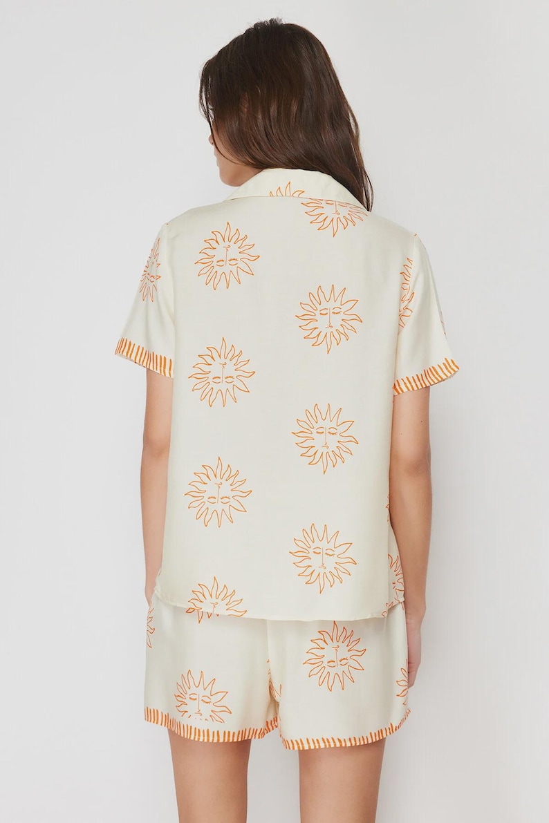 Op de afbeelding: Cr&egrave;mekleurige pyjamaset met een herhaald oranje zonnegroetpatroon. De top met korte mouwen en bijpassende shorts hebben een oranje rand. De set is gemaakt van een zachte, lichtgewicht stof.