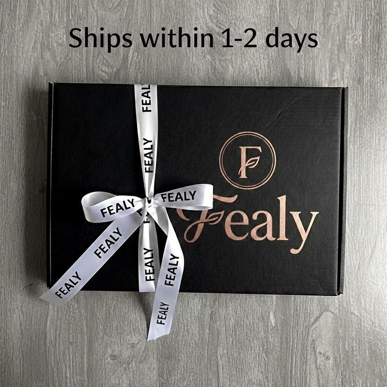 Op de afbeelding: Een zwarte geschenkdoos vastgebonden met een wit lint bedrukt met "FEALY". De doos heeft een gouden logo en het woord "Fealy" in gouden script. De tekst "Ships within 1-2 days" is zichtbaar.