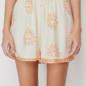 Op de afbeelding: Cr&egrave;mekleurige shorts met een elastische tailleband en een herhalend oranje zonontwerp. De shorts hebben een oranje gestreepte rand aan de zoom. De shorts maken deel uit van een bijpassende set.