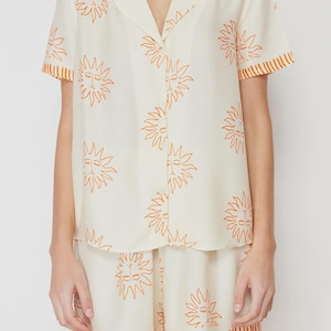 Op de afbeelding: Cr&egrave;mekleurige pyjamaset met een herhalend oranje zonontwerp. De set bevat een button-down shirt met korte mouwen en een kraag en bijpassende shorts. De randen van de mouwen en shorts zijn afgewerkt met oranje strepen.