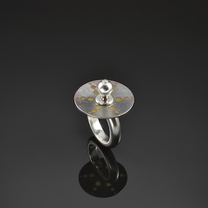 May include: A silver ring with a round, textured top featuring a circular design in shades of gold and copper. The top of the ring has a small, round knob.