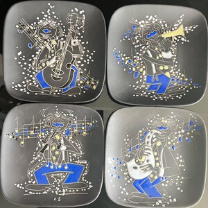 May include: Set of four square black ceramic plates with musical instrument designs. Each plate features a musician in blue and white, playing a different instrument: guitar, trumpet, flute, and saxophone. White musical notes and accents surround the figures.