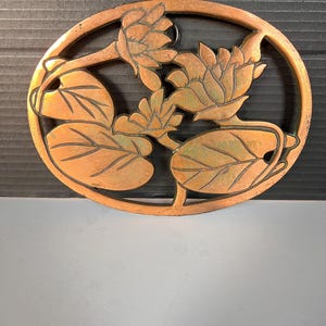 May include: An oval-shaped copper-toned metal trivet featuring an openwork design of two blooming lotus flowers and large lily pad leaves. The surface displays a weathered patina with dark recessed lines highlighting the floral details.