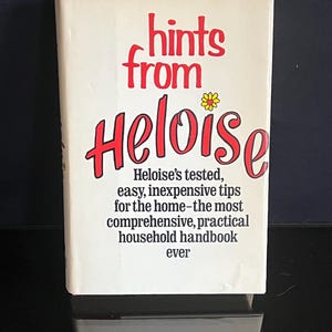 May include: A vintage book titled "Hints from Heloise" with red lettering and a small yellow flower. The book cover is off-white with text describing household tips. The book is on a reflective surface.