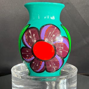 May include: A hand-painted ceramic vase in a vibrant teal color. The vase features a large, stylized flower design with purple petals, a red center, and green accents. The vase is displayed on a clear glass stand.