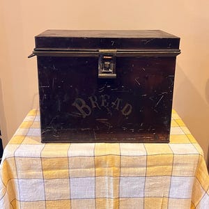 May include: A rectangular dark brown metal bread box featuring a hinged lid with a central metal latch. The front surface displays the word BREAD in a weathered, stenciled font. The exterior finish shows visible scratches and signs of wear.