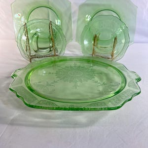 May include: A collection of green Depression glass tableware. The set includes square plates, small round bowls, and a large oval serving platter. The glass has a delicate floral pattern and a scalloped edge. The pieces are arranged on a white surface.