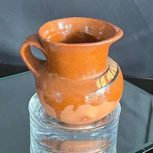 May include: A small, brown ceramic pitcher with a handle and a wide opening. The pitcher has a decorative band of black and gold near the top. It sits on a clear, glass base.