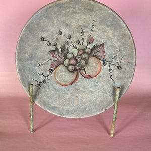 May include: A round, hand-painted plate with a fruit design, including grapes and peaches. The plate has a gray background and is displayed on a metal stand. The fruit is painted in shades of red, green, and brown.
