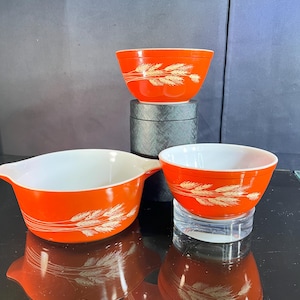 May include: A set of vintage Pyrex bowls in a vibrant orange hue, featuring a wheat pattern in cream. The bowls have white interiors and are arranged in a nested display. The set includes a bowl with handles.