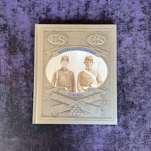 May include: A book titled "The Civil War Brother Against Brother" with images of soldiers. The cover features the letters "US" and "CS" with crossed cannons and the words "The War Begins". The book is on a purple surface.