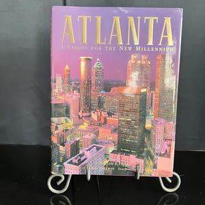 May include: A book titled "ATLANTA A Vision for the New Millennium" with a cityscape image on the cover. The cover is purple with gold lettering. The book is displayed on a decorative metal stand.