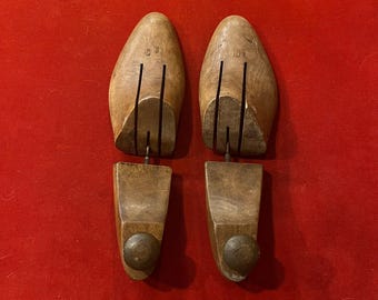 May include: Two wooden shoe trees, each with a rounded toe shape and a heel piece. The toe section has vertical grooves and the heel piece has a metal knob. The shoe trees are displayed on a red surface.