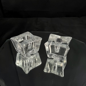 May include: Two clear glass star-shaped candle holders. Each holder has a central circular opening for a candle. The holders are on a reflective surface, creating a mirrored effect. The background is dark.