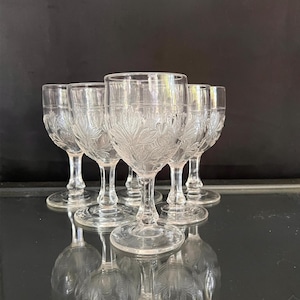 May include: A set of six clear glass goblets featuring a delicate embossed floral pattern on the bowl. Each goblet has a smooth, rounded rim, a short stem, and a circular base, reflecting light across the surface of the clear glass material.