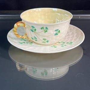 May include: Cream-colored teacup and saucer set with a shamrock design. The teacup has a yellow interior and a handle with orange and green accents. The saucer features a woven pattern. The set is ideal for serving tea.