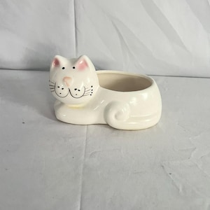 May include: A white ceramic cat-shaped planter with a smiling face, pink ears, and black whiskers. The planter has an open top for holding small plants or items. The cat is in a lying down position.