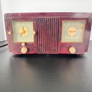 May include: A vintage General Electric radio clock with a dark red marbled plastic casing. The device features a square analog clock face on the left and a radio frequency dial on the right, separated by a vertical slatted speaker grille.
