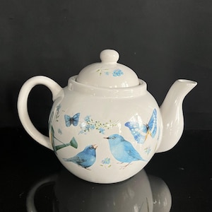 May include: White ceramic teapot with a lid, handle, and spout. The teapot is decorated with blue birds, butterflies, and floral accents. The teapot is sitting on a reflective surface.