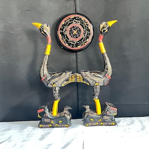 May include: Two decorative black and yellow ostrich sculptures with intricate patterns. The ostriches have long necks and are mounted on decorative bases. A round, black and red wall hanging with a gold design is centered above the sculptures.