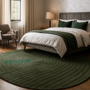 May include: A dark green, circular rug with a concentric spiral design sits on a hardwood floor in a bedroom. The rug is in front of a bed with white bedding and green pillows and a green blanket. The text "shama carpet" is visible on the rug.