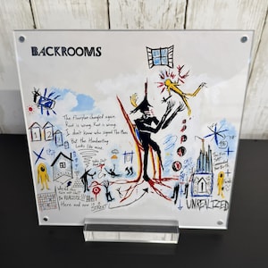 May include: A square acrylic display featuring a white background with colorful, chaotic hand-drawn illustrations and text including BACKROOMS, The Floorplan changed again, and UNREALIZED. The artwork depicts abstract figures, buildings, and symbols.