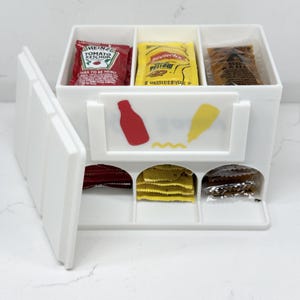 Puede incluir: Un organizador de condimentos de plástico blanco con tres compartimentos verticales y una puerta lateral abatible. El panel frontal presenta iconos con forma de botella en rojo y amarillo. Contiene sobres individuales de kétchup, mostaza y salsa picante.