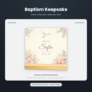 May include: A square plexiglass baptism keepsake print featuring a watercolor dove, pink flowers, and green foliage. The text reads "child of God", "Sofia", "BAPTIZED", and "May 17, 2026". The print is displayed on a rectangular light-colored beechwood base.