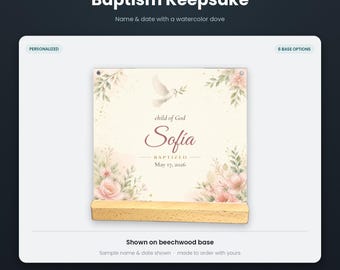 May include: A square plexiglass baptism keepsake print featuring a watercolor dove, pink flowers, and green foliage. The text reads "child of God", "Sofia", "BAPTIZED", and "May 17, 2026". The print is displayed on a rectangular light-colored beechwood base.