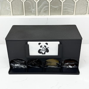Puede incluir: Un organizador de almacenamiento rectangular negro con cuatro compartimentos inferiores que contienen pequeños paquetes de aluminio. Una etiqueta rectangular blanca está fijada al frente, con una ilustración gráfica negra de un panda comiendo fideos.