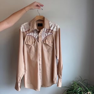 Op de afbeelding: Een beige overhemd met lange mouwen en een western-stijl ontwerp. De blouse heeft witte kanten details op de schouders en het juk, met bordeauxrode stiksels. Het heeft twee borstzakken en manchetten met knopen.