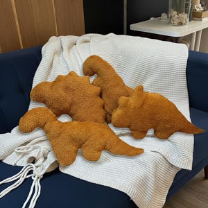 May include: Four plush dinosaur-shaped pillows in varying designs and sizes. The pillows are a warm, golden-brown color and rest on a white knitted blanket draped over a dark blue sofa. The pillows are soft and textured.