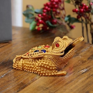 Può includere: Statuetta a forma di rana dorata con dettagli di gioielli rossi, blu e verdi. La rana tiene una moneta in bocca. Poggia su una superficie di legno, con uno sfondo sfocato di bacche rosse.