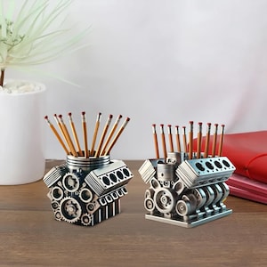Può includere: Due organizer da scrivania color argento a forma di motori V8, ciascuno con matite. Gli organizer presentano dettagliati disegni di ingranaggi e componenti del motore. Uno è leggermente angolato, mostrando l'intricata meccanica.