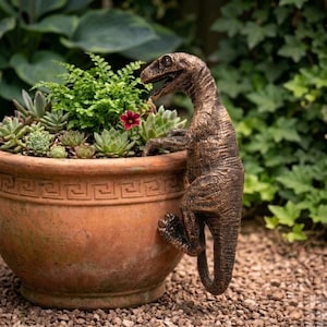 May include: A bronze dinosaur sculpture clings to a terracotta plant pot filled with succulents and greenery. The pot has a decorative border and sits on a bed of small pebbles, with a lush green background.