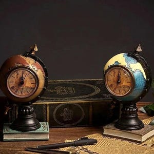 May include: Two globe-shaped clocks with antique styling. One clock features brown and gold tones, the other blue and yellow. Both have a black base and a clock face with Roman numerals. They sit on small books.