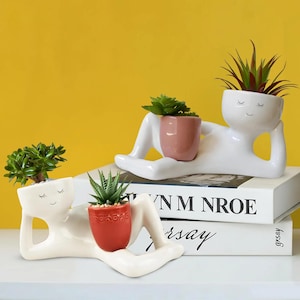 May include: Two white ceramic planters shaped like reclining figures, each holding a small potted succulent. The planters have a minimalist design with closed eyes and a smiling face. One holds a red pot, the other a pink pot.