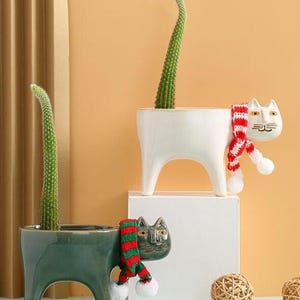 May include: Two cat-shaped ceramic planters, one white and one dark green, each holding a tall, green cactus. The white cat wears a red and white striped scarf. The green cat has a red and green scarf. Decorative home decor.