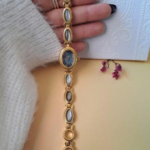 Op de afbeelding: Een goudkleurig schakelhorloge met een ovale wijzerplaat in blauw met Arabische cijfers. De armband bestaat uit afwisselende ovale iriserende cabochons en gestructureerde goudkleurige metalen schakels die een decoratieve ketting vormen.