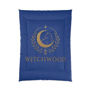 May include: A blue comforter featuring a gold design with a crescent moon, cat silhouette, star, and the word "WITCHWOOD". The design is surrounded by a laurel wreath and a lantern.
