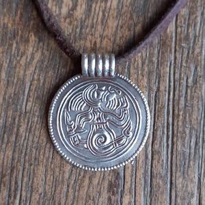 May include: A silver pendant with an intricate design, suspended from a brown suede cord. The pendant features a detailed, embossed pattern within a circular frame. The cord is a dark brown color, and the pendant is made of a shiny metal.