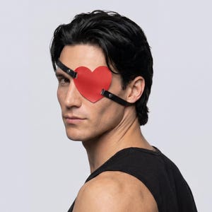 May include: A red heart-shaped eye patch with black straps. The eye patch is made of a smooth material and is worn over one eye. The straps are secured with small metal rivets. The person is wearing a black tank top.