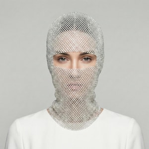 Puede incluir: Un pasamontañas de malla plateada cubre la cabeza y el cuello, con aberturas para los ojos y la boca. El pasamontañas está hecho de pequeños cuadrados reflectantes, creando un efecto brillante. La persona lleva una camiseta blanca.