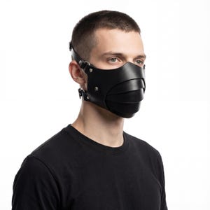 Pode incluir: Uma máscara facial de couro preto com um design em camadas. A máscara tem alças ajustáveis com ferragens de metal e cobre o nariz e a boca. A máscara é usada por uma pessoa com uma camiseta preta.