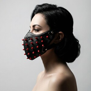 May include: Black leather face mask with red spikes. The mask has a contoured shape and is secured with adjustable straps. The red spikes are evenly spaced across the front of the mask, creating a bold and edgy aesthetic.