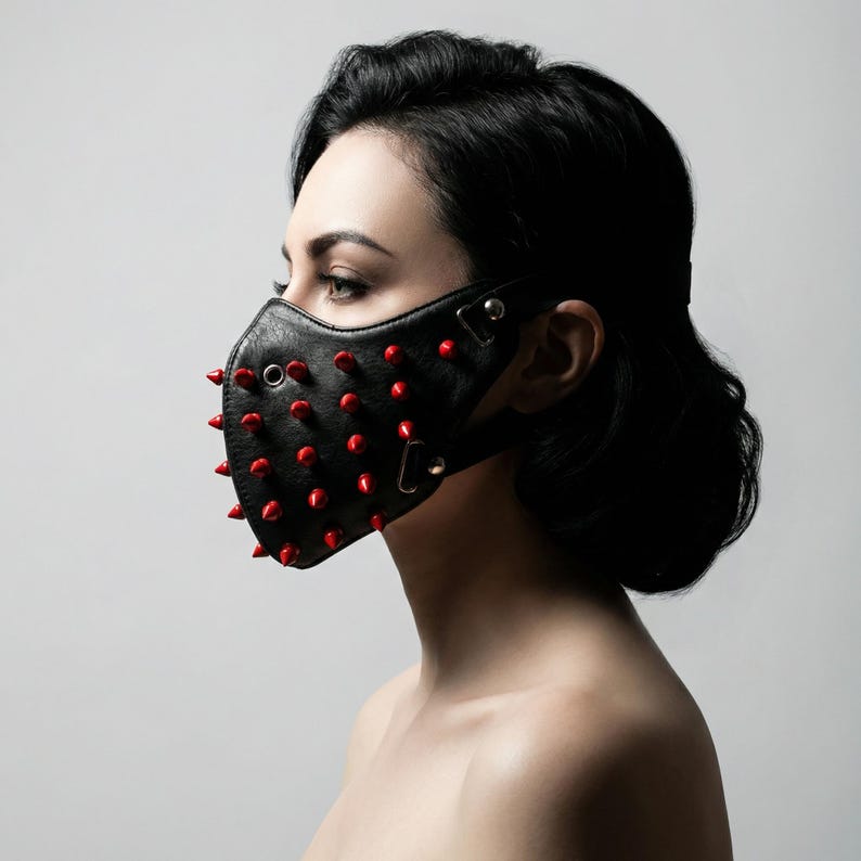 May include: Black leather face mask with red spikes. The mask has a contoured shape and is secured with adjustable straps. The red spikes are evenly spaced across the front of the mask, creating a bold and edgy aesthetic.