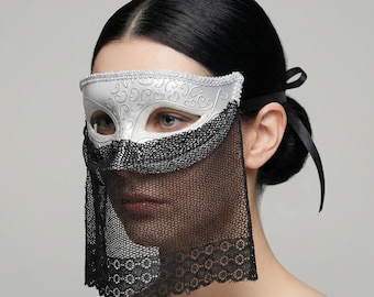 May include: A white and silver masquerade mask with a black lace veil. The mask has a decorative floral design and is secured with a black ribbon. The mask covers the eyes and nose.