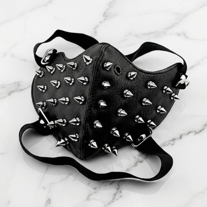 May include: Black leather face mask with numerous silver-colored cone studs. The mask has black straps and metal hardware for attachment. The mask is designed to cover the nose and mouth.