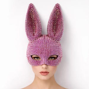 Puede incluir: Una máscara de conejo rosa enjoyada con largas orejas. La máscara cubre los ojos y la nariz, con aberturas para los ojos. La máscara está cubierta de pequeñas gemas rosas brillantes. El fondo es blanco liso.