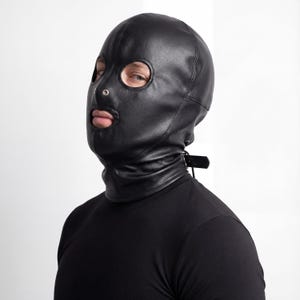 Może przedstawiać: Czarny skórzany kaptur z otworami na oczy i usta. Kaptur zakrywa całą głowę i szyję, z małym metalowym pierścieniem na nosie. Pasek z klamrą jest widoczny z tyłu szyi.